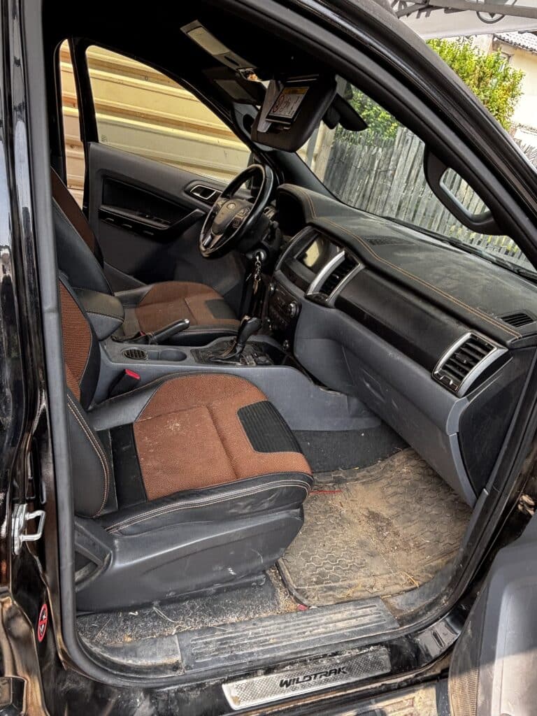 Intérieur Ford Ranger Wildtrak côté conducteur, très sale et poussiéreux. Sièges noir et orange, tapis boueux, texte WILDTRAK visible.