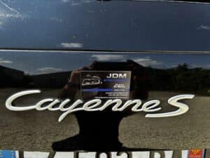 Emblème "Cayenne S" sur carrosserie noire rayée. Réflexion du photographe et carte de visite "JDM Auto Cleaning" (Jérémy De Matos).