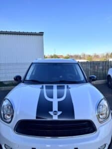 MINI Countryman blanche, vue de face. Bandes de rallye noires stylisées sur le capot. Logo MINI et phares ronds visibles sous ciel bleu.