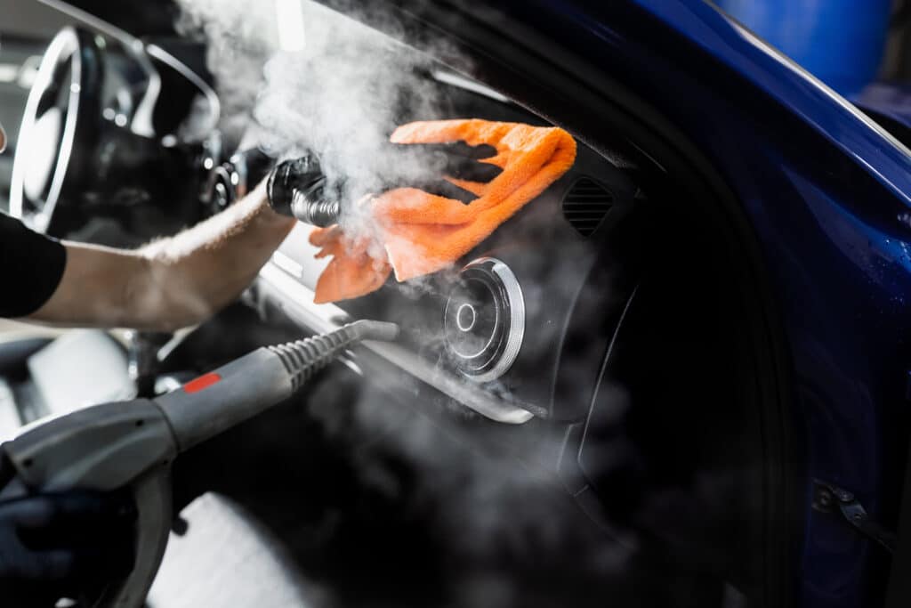Nettoyage vapeur voiture intérieur automobile Nettoyage intérieur de voiture à la vapeur : un technicien désinfecte le tableau de bord avec un pistolet à vapeur et un chiffon orange.