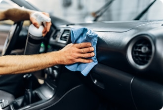Nettoyage intérieur voiture : désinfection et soin tableau de bord Homme nettoyant le tableau de bord d'une voiture avec un chiffon bleu et un vaporisateur de produit nettoyant pour intérieur automobile.