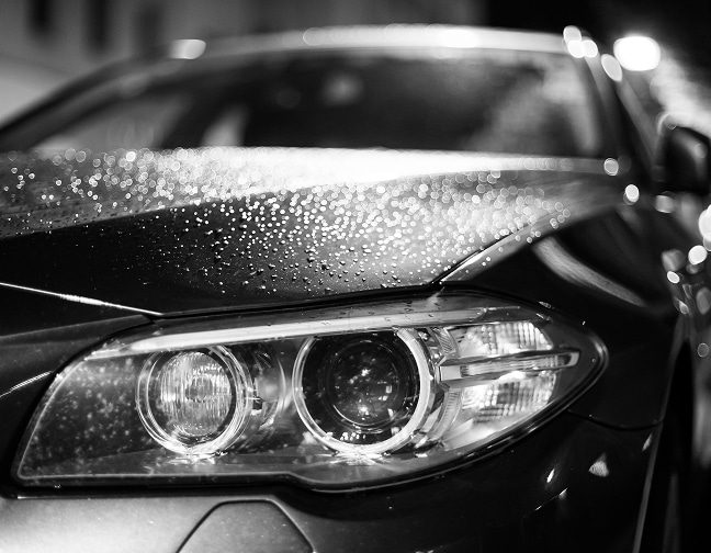 BMW de nuit sous la pluie : gros plan phare et gouttes Gros plan nocturne en noir et blanc sur le phare avant d'une BMW avec des gouttes de pluie formant des bokeh lumineux sur le capot.