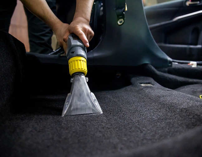 Nettoyage Inject-Extract Tapis Voiture Professionnel Détail gros plan d'un professionnel nettoyant en profondeur la moquette noire d'une voiture avec un injecteur-extracteur professionnel muni d'une buse transparente.
