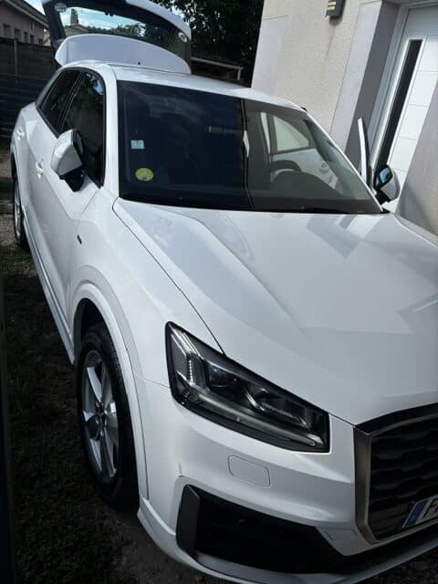 Audi Q2 SUV blanc : Hayon et porte ouverts après le nettoyage. Audi Q2 blanc, hayon et porte ouverts, garé devant une maison. Détails des phares LED, jantes alliage et vignette jaune sur le pare-brise.