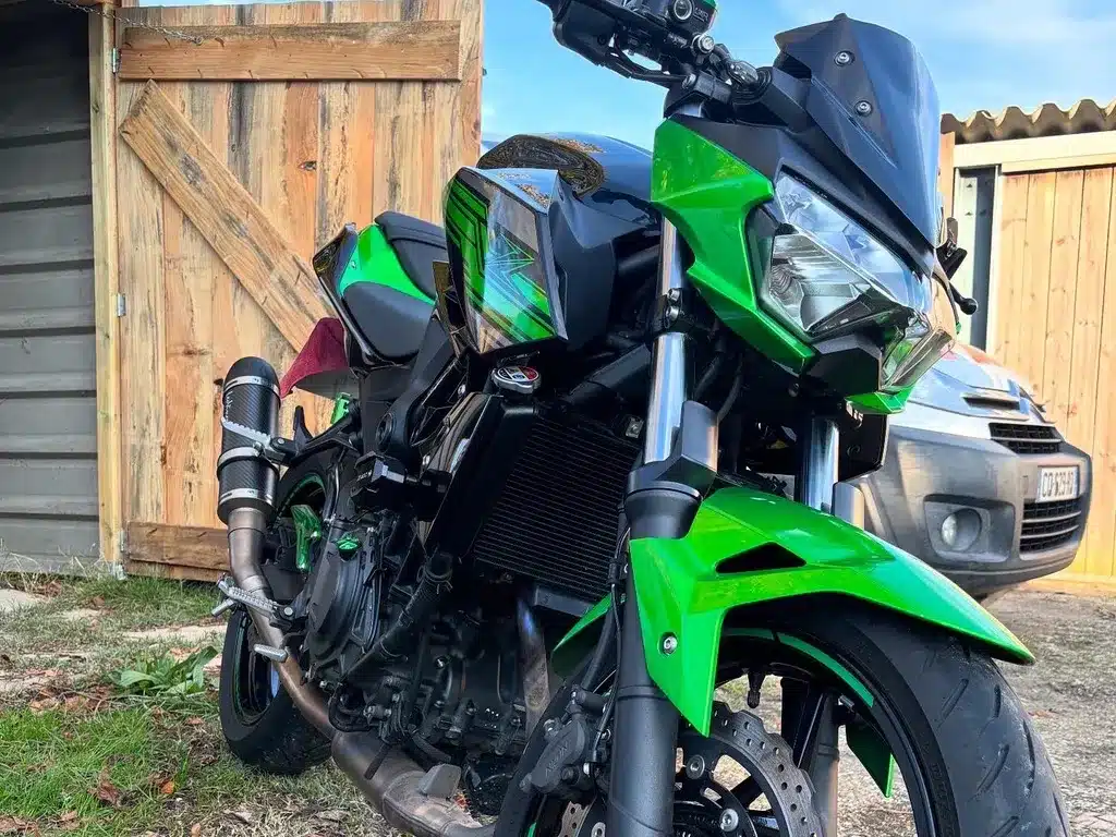 Kawasaki Z vert : La moto naked la plus agressive Moto naked vert Kawasaki et noir, vue avant. Échappement carbone, radiateur exposé et carénage agressif devant une porte en bois rustique.