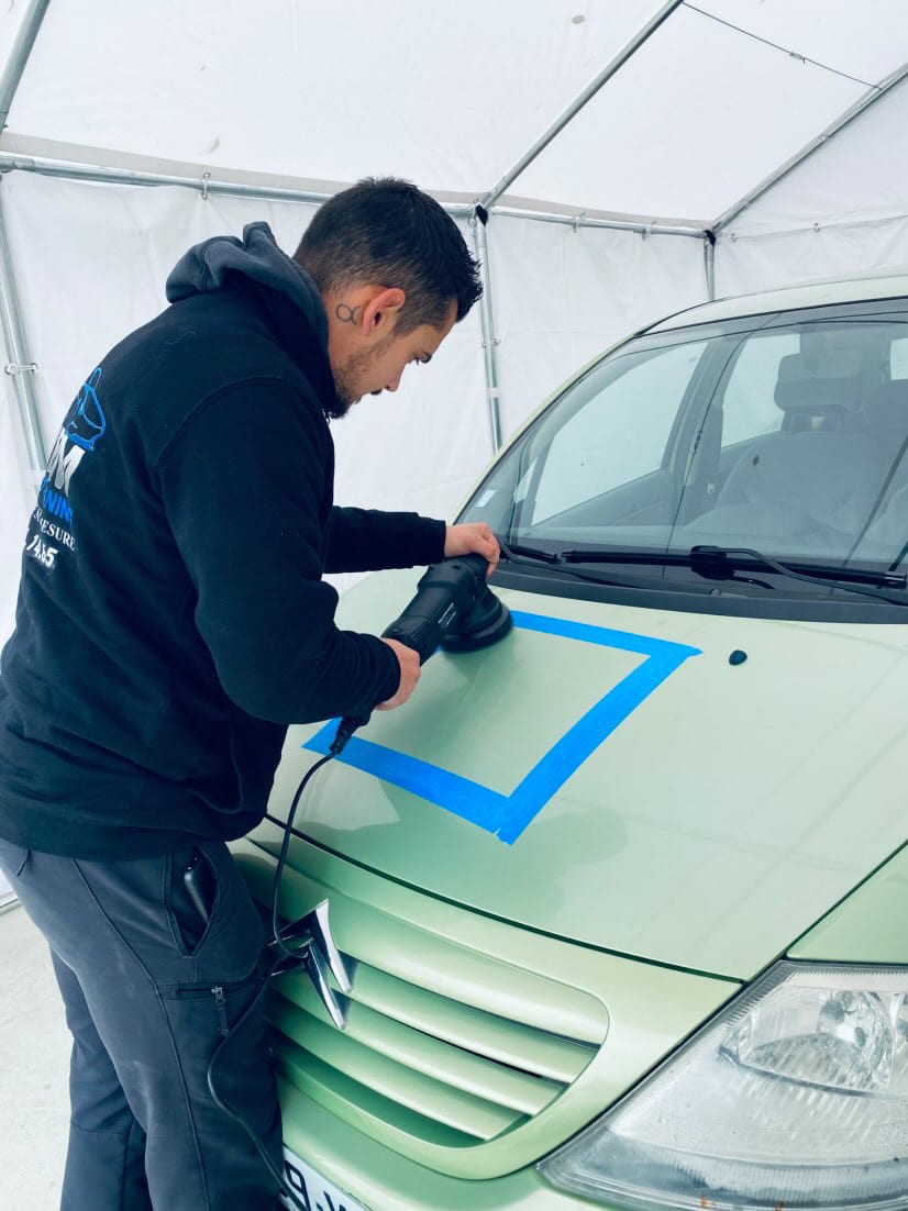 Detailing Auto : Polissage de carrosserie et test spot. Professionnel du detailing effectuant un polissage de la carrosserie. Polisseuse orbitale sur capot vert délimité par ruban bleu.