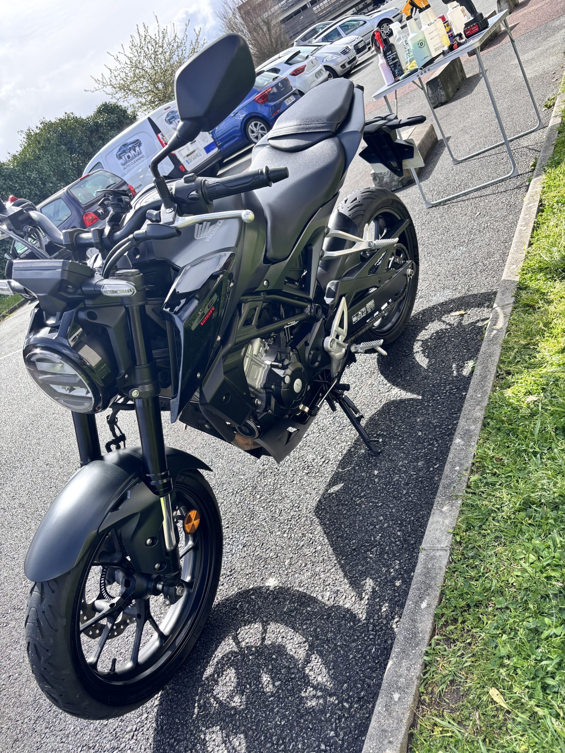 Moto Honda CB125R noire stationnée devant un stand de nettoyage JDM Auto Cleaning avec produits d'entretien.