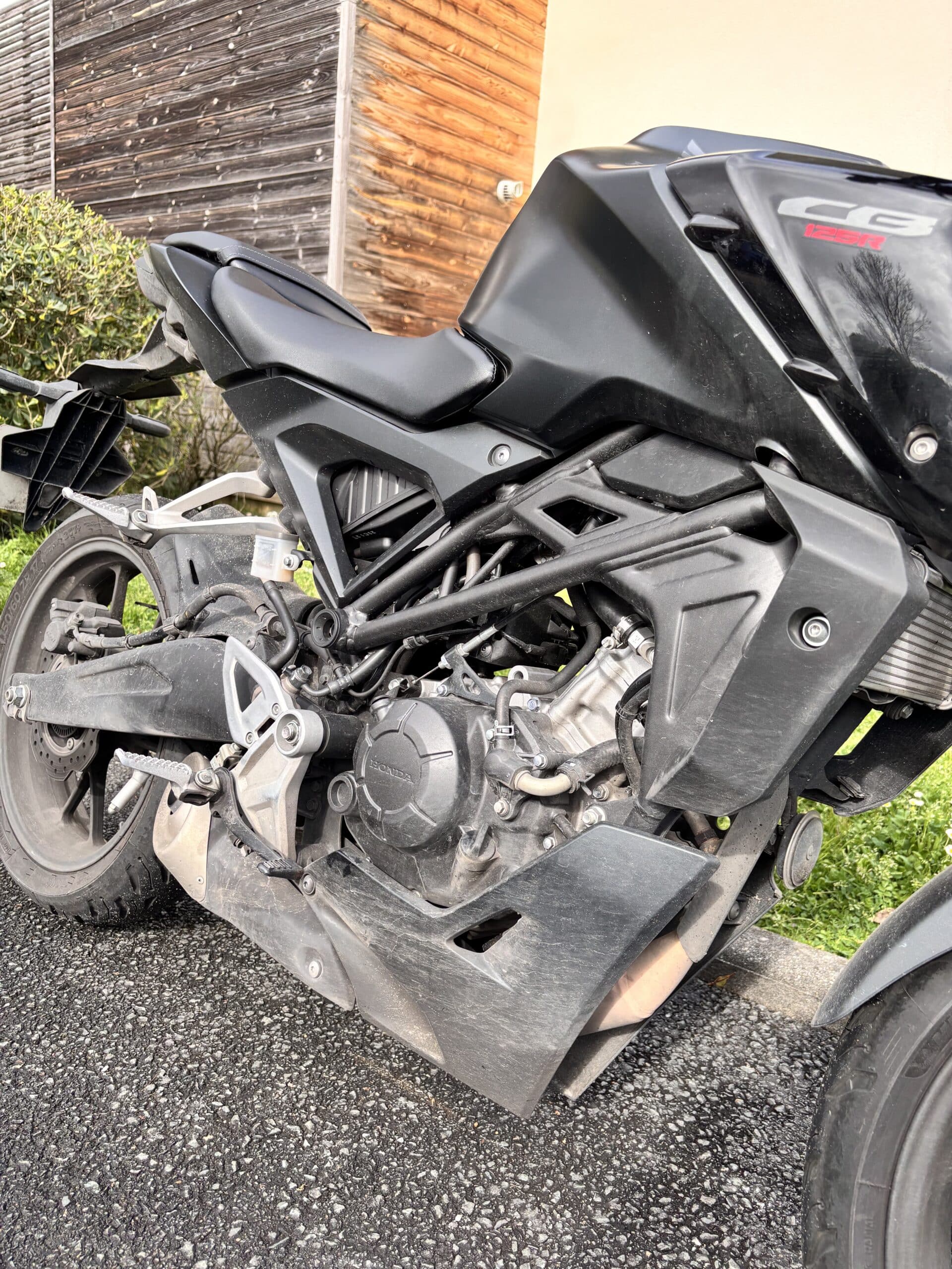 Moto Honda CB125R noire : gros plan sur le moteur, le cadre en treillis et la roue arrière sur bitume.