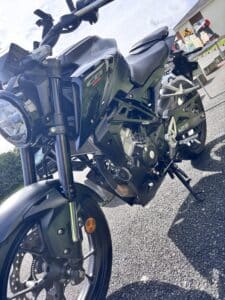 Moto Honda CB125R noire avec phare LED et cadre apparent, stationnée sur asphalte par une journée ensoleillée.