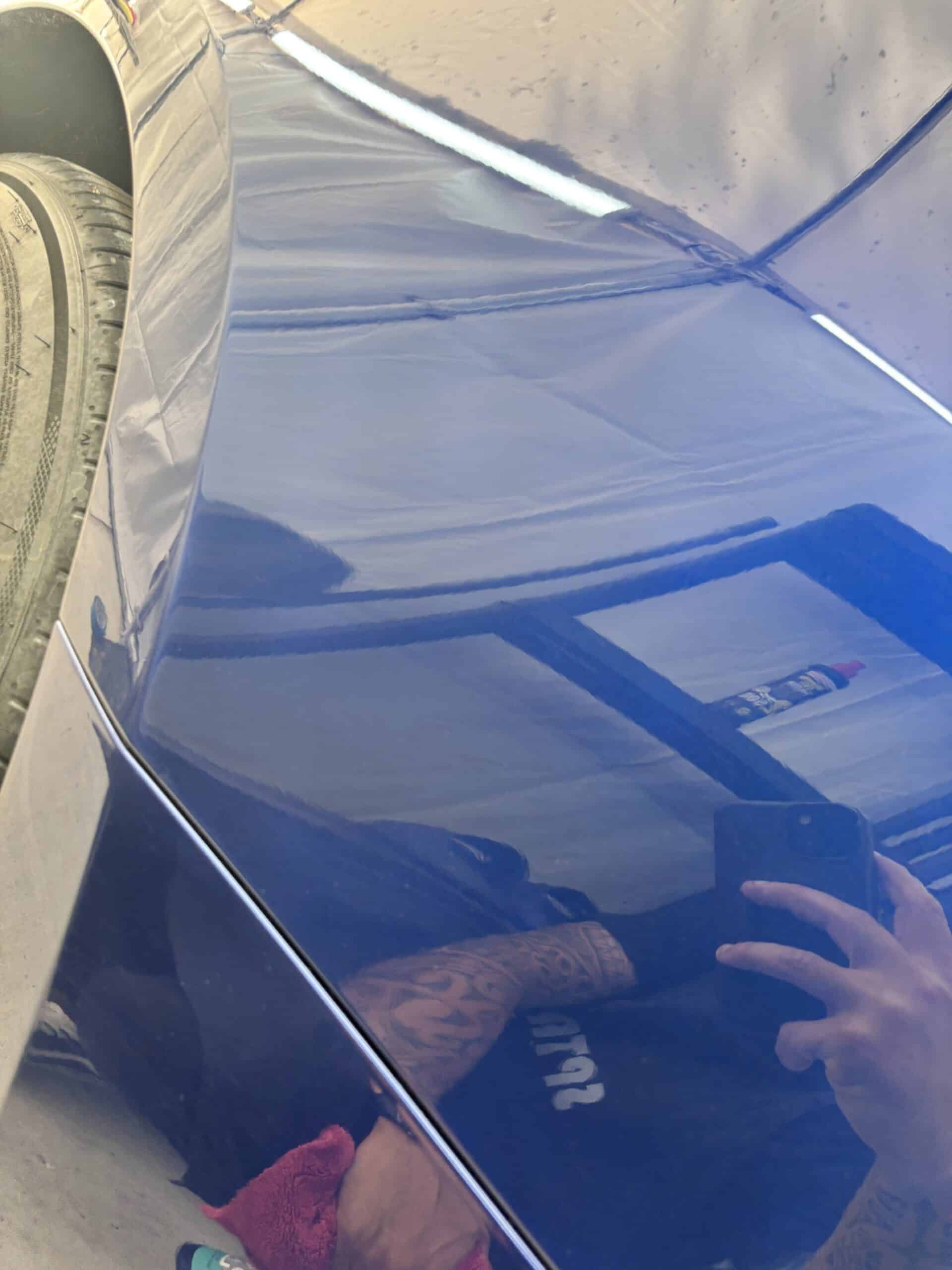 Détail de carrosserie bleue avec reflet d'un homme tatoué prenant une photo, résultat d'un polissage auto.