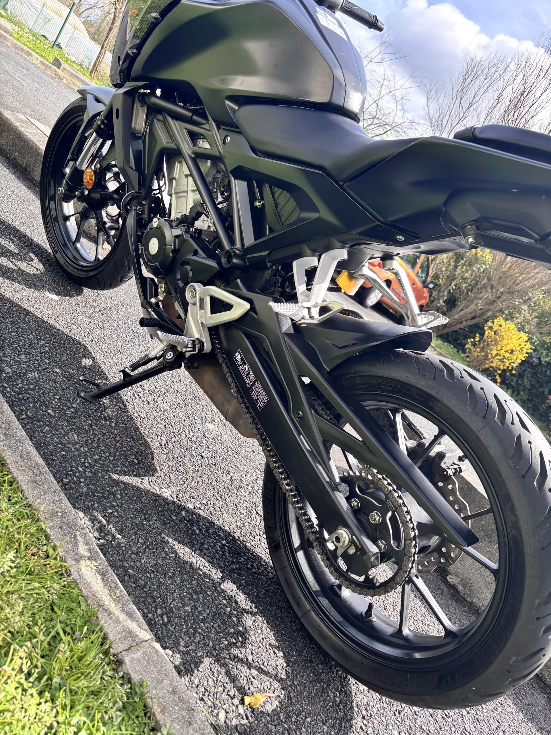 Moto noire Honda CB garée, focus sur la chaîne et la roue arrière, pneu Michelin, par temps ensoleillé.