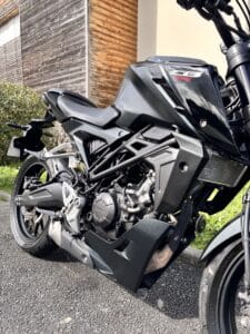 Moto Honda CB125R noire stationnée montrant son cadre et moteur devant un bâtiment en bois.