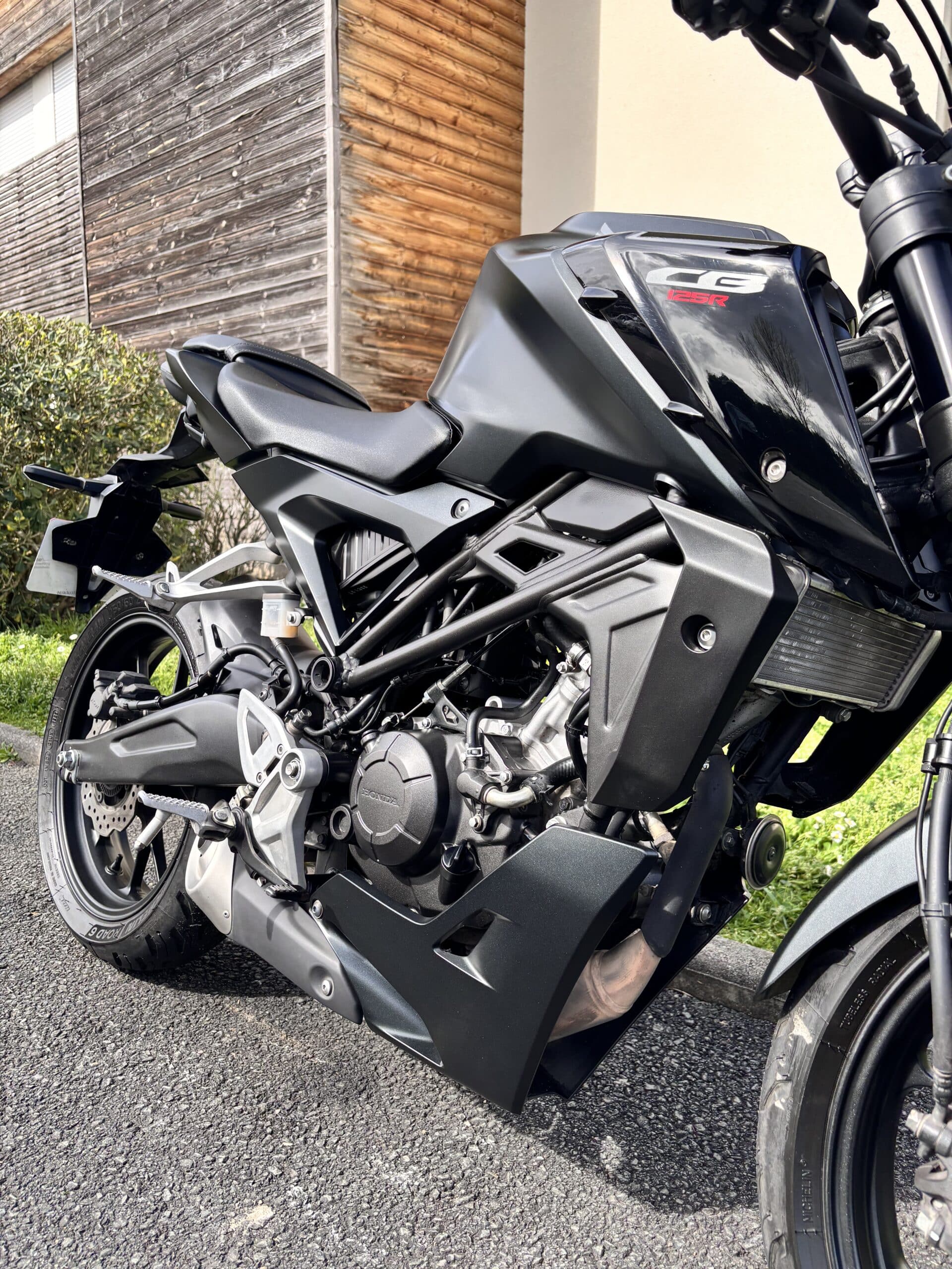 Moto Honda CB125R noire stationnée montrant son cadre et moteur devant un bâtiment en bois.