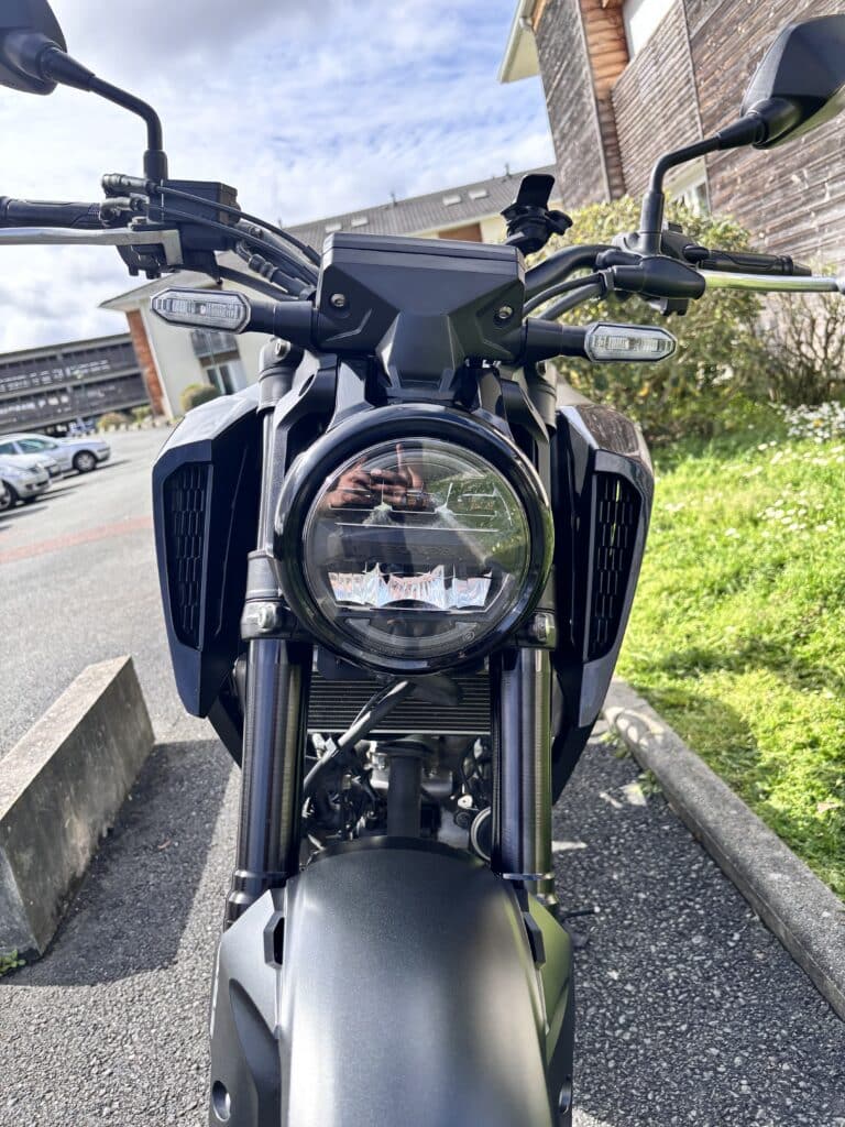Vue avant d'une moto Honda noire avec phare LED rond distinctif, stationnée devant des bâtiments.