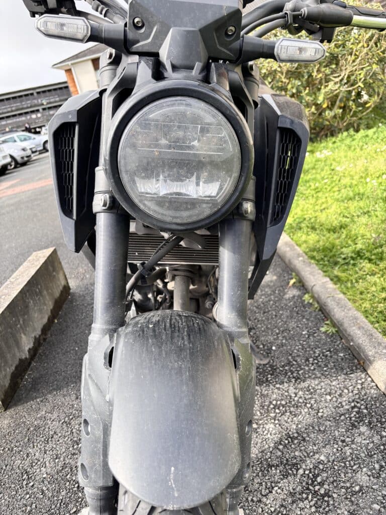 Phare LED rond et fourche d'une moto noire moderne sur un parking, garde-boue avant poussiéreux.