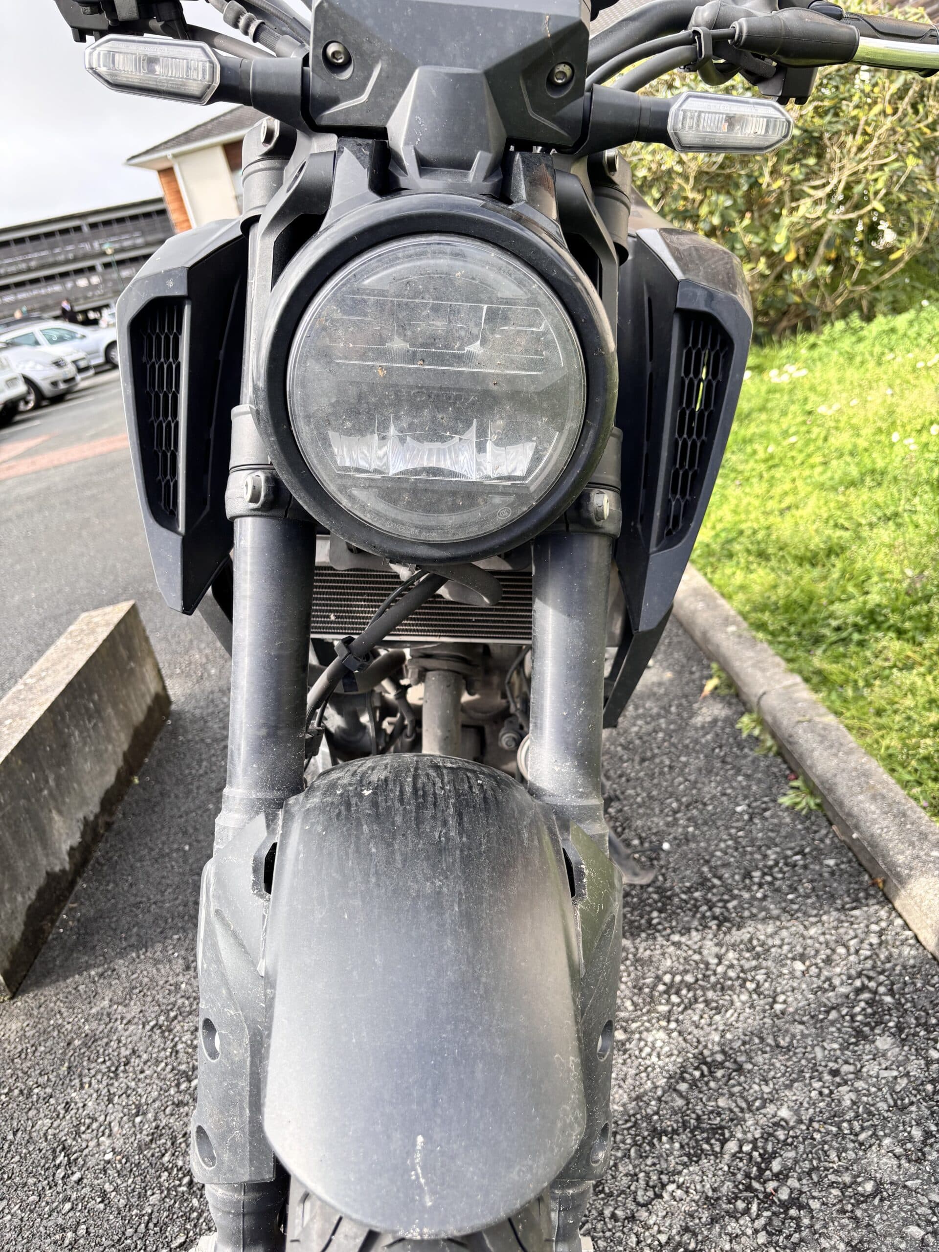 Phare LED rond et fourche d'une moto noire moderne sur un parking, garde-boue avant poussiéreux.
