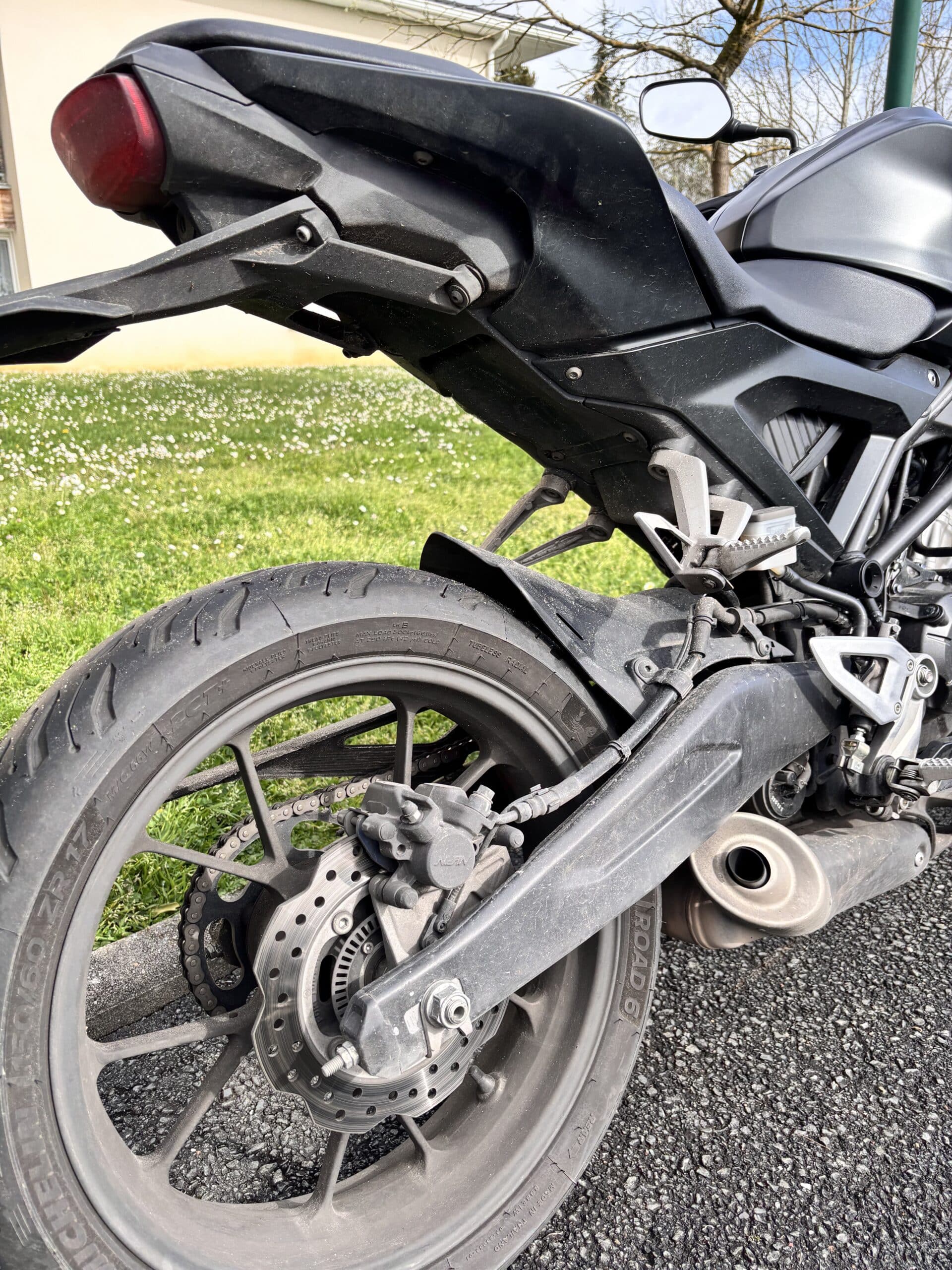 Roue arrière de moto avec pneu Michelin Road 6, frein à disque et pot d'échappement sur fond de pelouse verte.