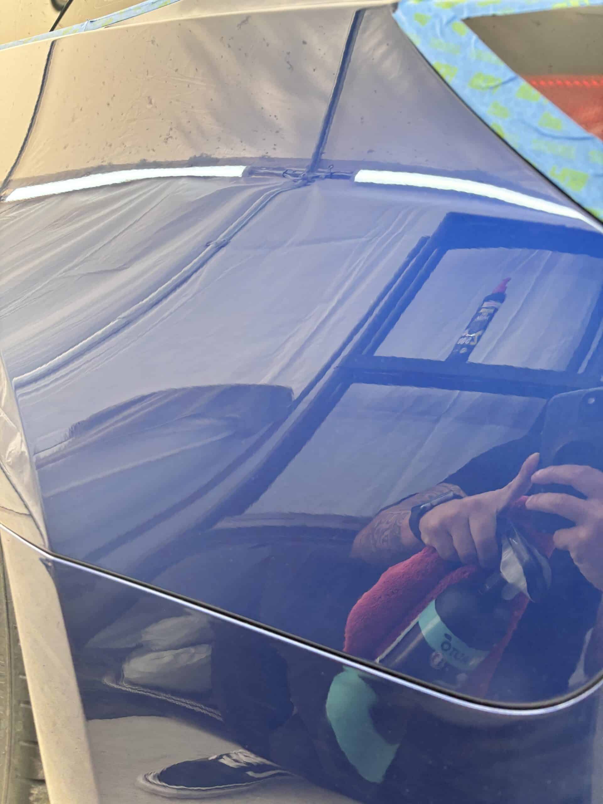 Reflet d'un technicien nettoyant une carrosserie bleue avec un spray et un chiffon microfibre rouge.